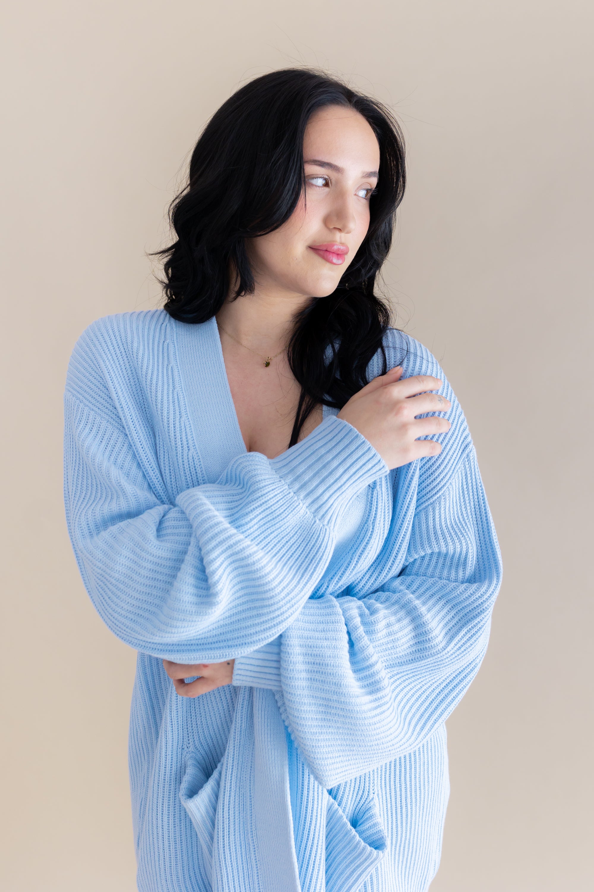 Female model wrapped in the Chunky Knit Women's Oversized Cardigan in Breeze standing in front of a light beige background