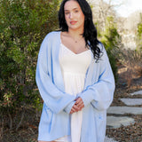Female model standing outside on a stone path wearing the Chunky Knit Women's Oversized Cardigan in Breeze with a light neutral colored dress on underneath