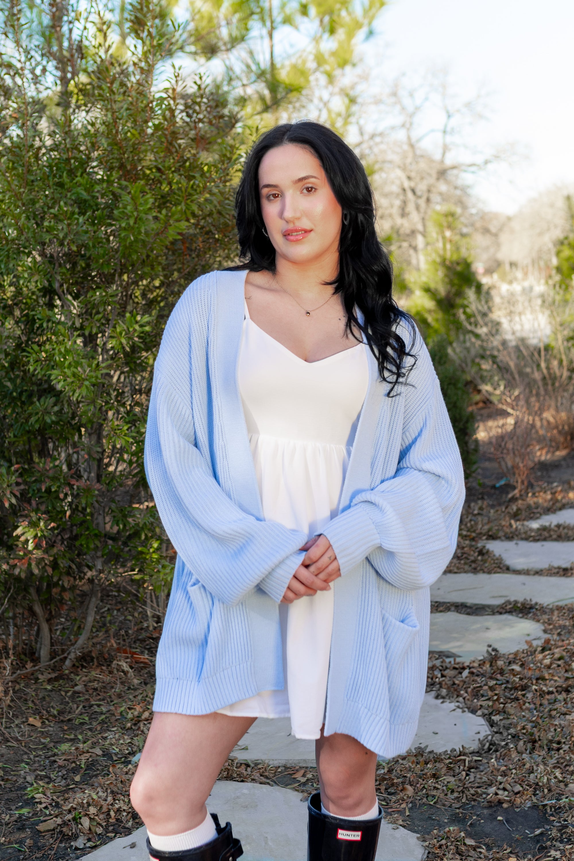Female model standing outside on a stone path wearing the Chunky Knit Women's Oversized Cardigan in Breeze with a light neutral colored dress on underneath