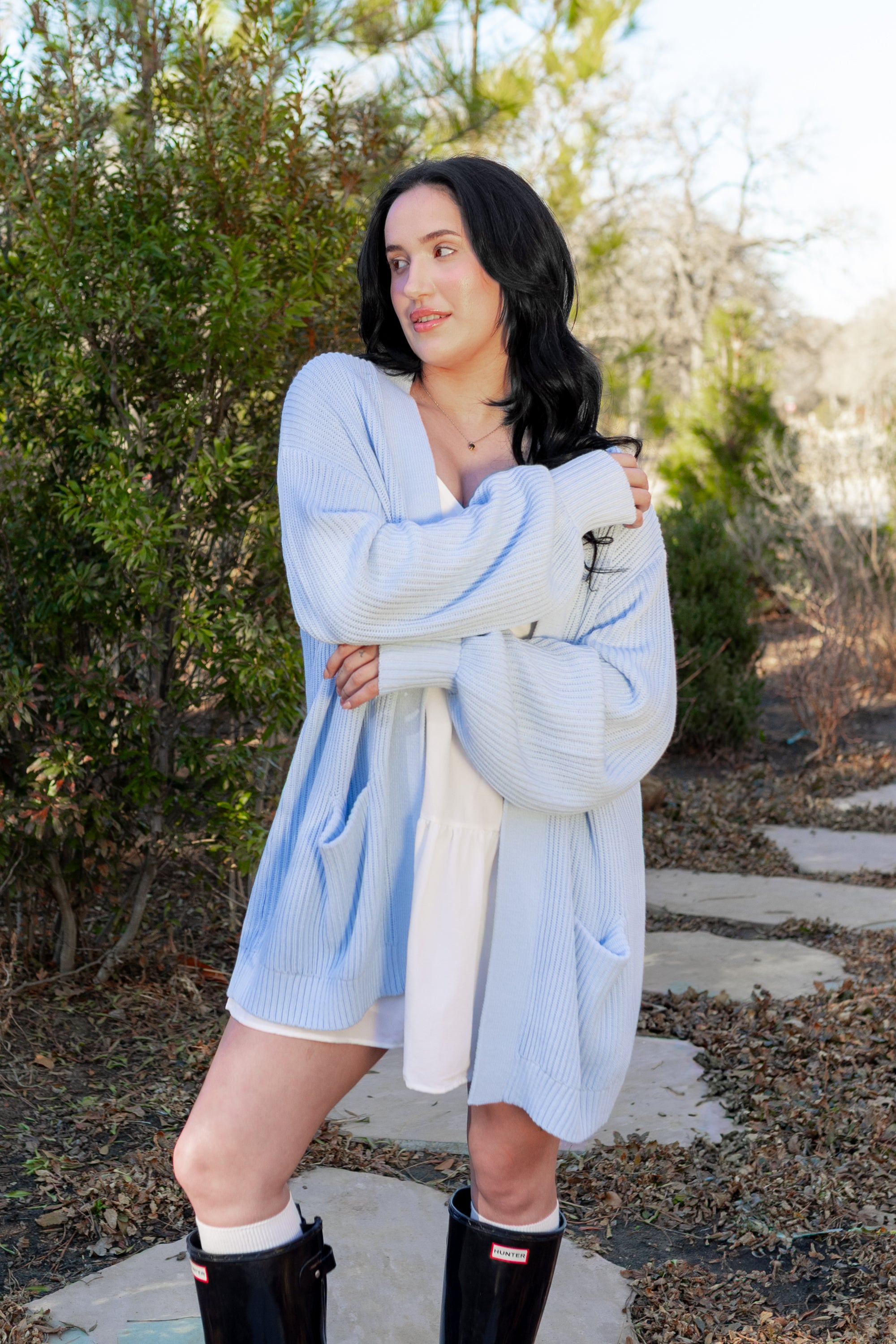 Female model standing outside on a stone path modeling the Chunky Knit Women's Oversized Cardigan in Breeze overtop