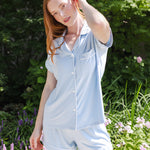 A woman wearing a light blue short-sleeve button-front pajama shirt and matching shorts, standing outdoors in a garden with green foliage and flowers in the background.
