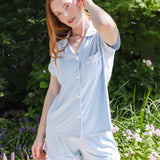 A woman wearing a light blue short-sleeve button-front pajama shirt and matching shorts, standing outdoors in a garden with green foliage and flowers in the background.