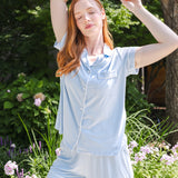 A woman posing outdoors in a light blue short-sleeve button-up pajama shirt and matching pajama shorts, set against a lush green garden background.