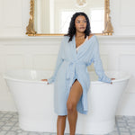 Woman in a light blue robe standing in a bathroom with white freestanding bathtub and gold-framed mirror.