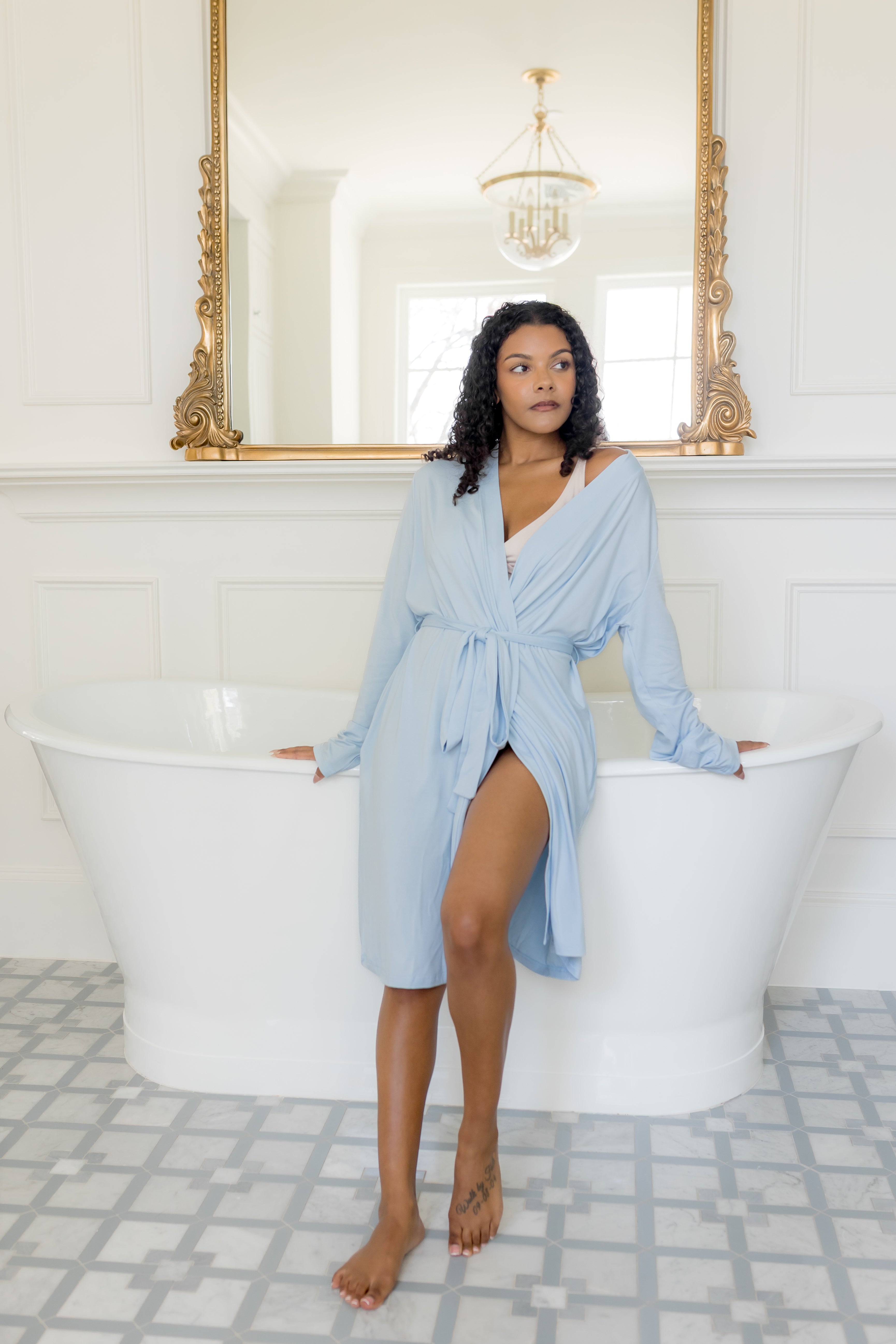 Woman in a light blue robe standing in a bathroom with white freestanding bathtub and gold-framed mirror.