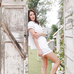 Woman in a white pajama standing in a rustic outdoor setting with wooden walls and greenery.