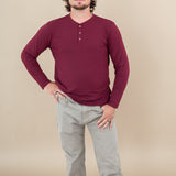 Male model with one hand on hips while wearing Kyte Long Sleeve Henley Tee in Burgundy paired with tan pants 