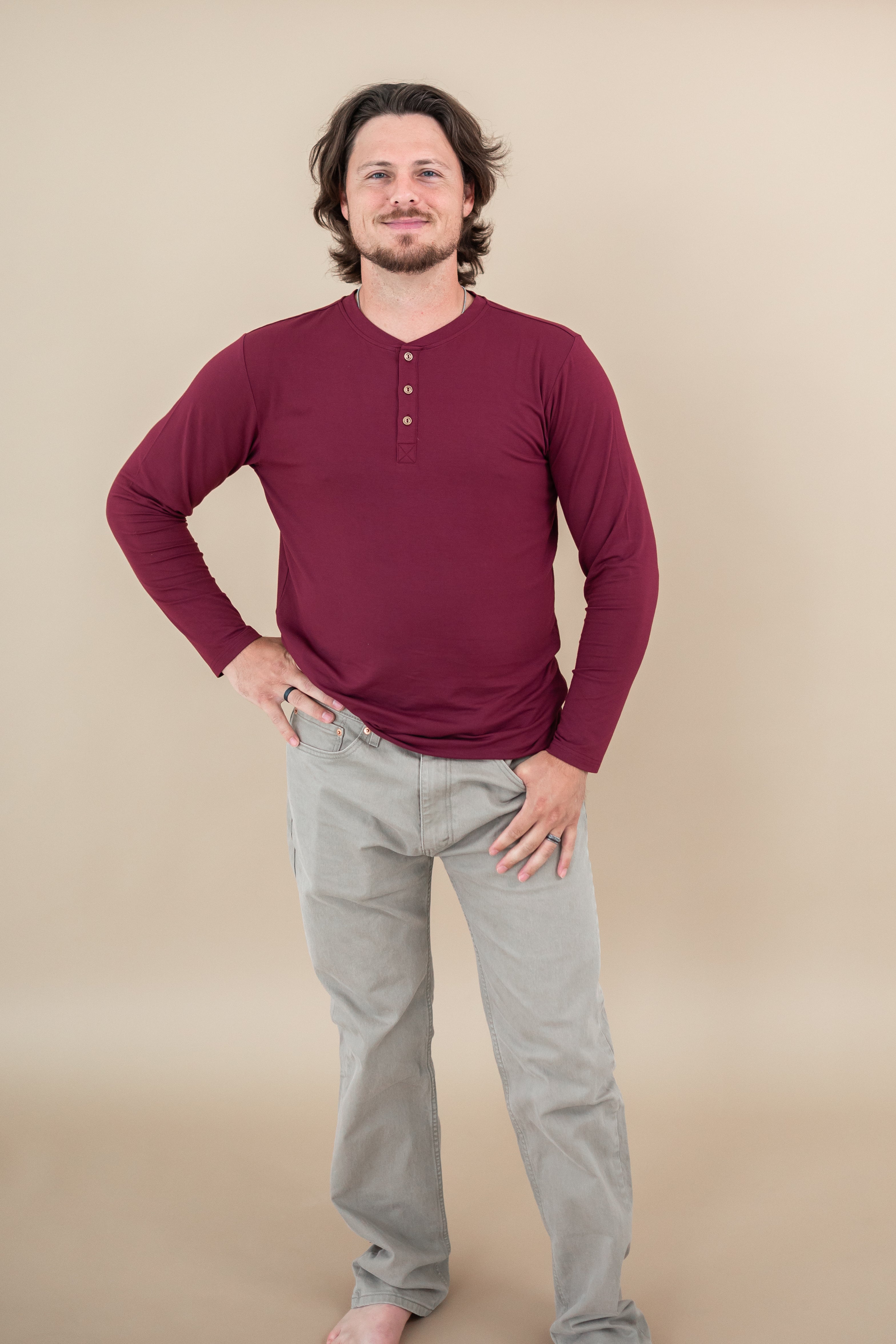 Male model with one hand on hips while wearing Kyte Long Sleeve Henley Tee in Burgundy paired with tan pants 