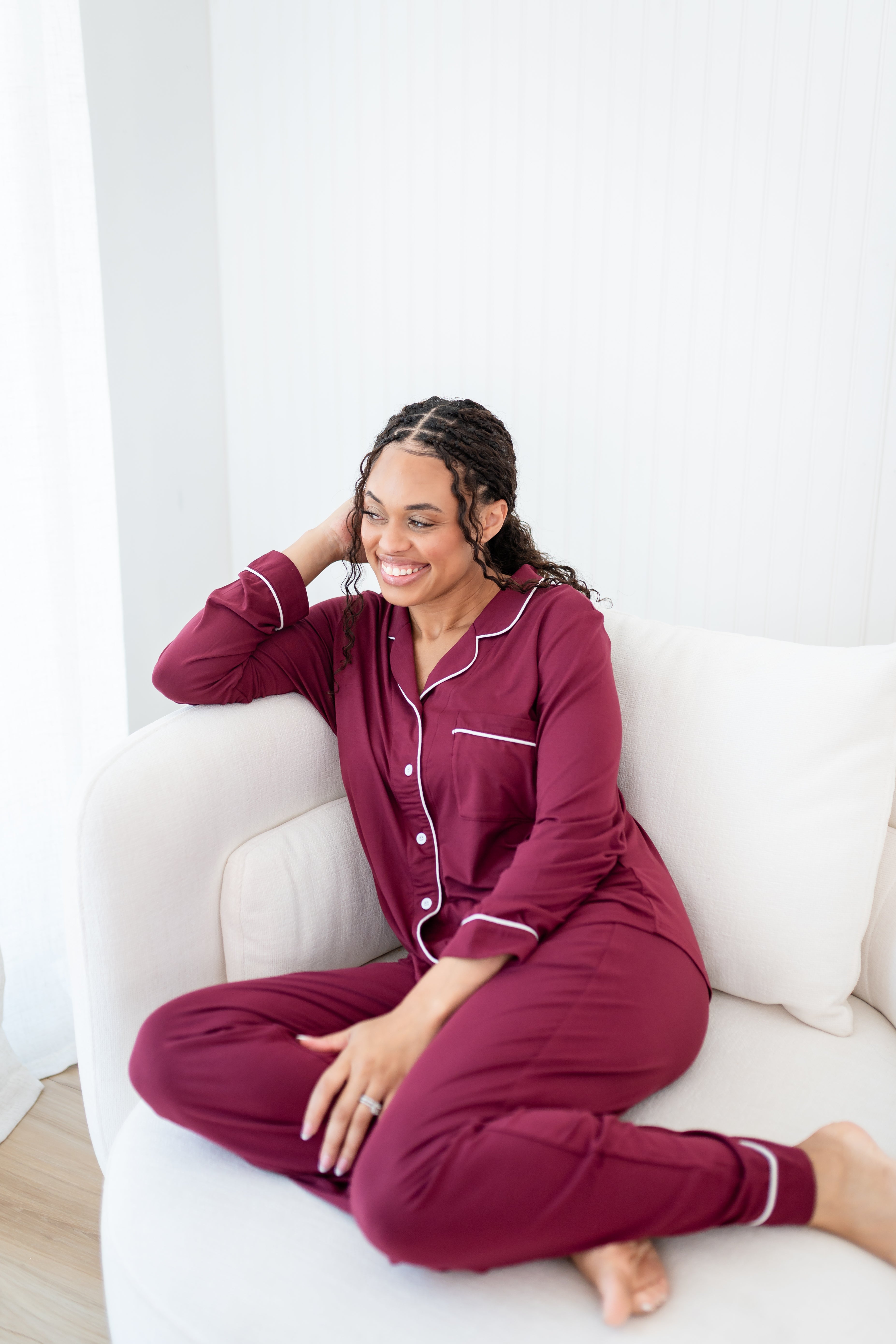 Smiling model sitting on a white couch while wearing Women's Long Sleeve Pajama Set in Burgundy