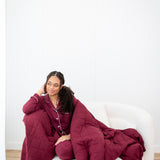 Model looking to the side while sitting on a white sofa chair wrapped in a Quilt Blanket in Burgundy 2.5 