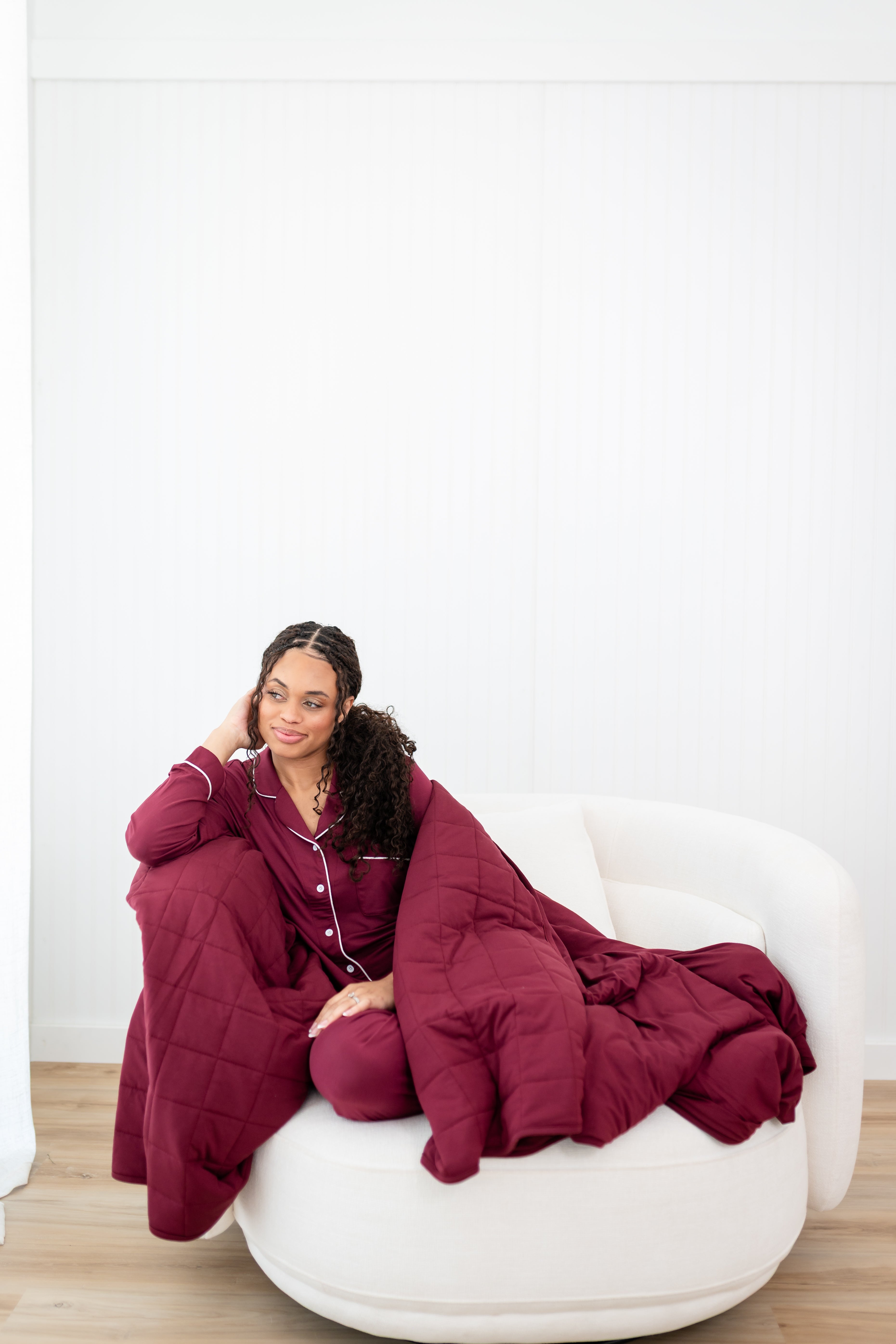 Model looking to the side while sitting on a white sofa chair wrapped in a Quilt Blanket in Burgundy 2.5 