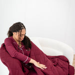 Model looking down at and wrapped in a Quilt Blanket in Burgundy 2.5 while wearing matching Long Sleeve Pajama Set