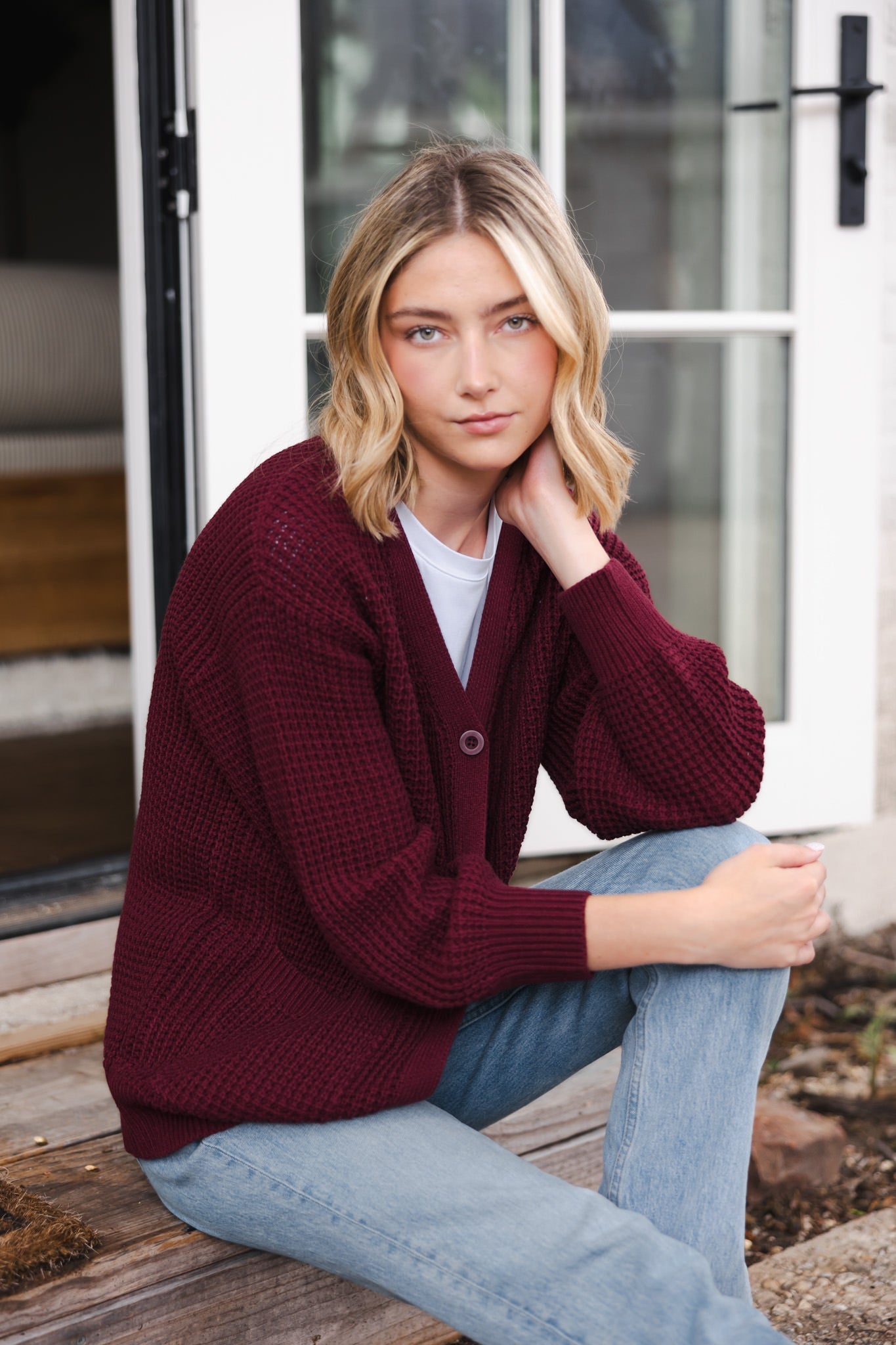 Modal sitting outdoors wearing Chunky Knit Women's Waffle Cardigan in Burgundy over white t-shirt and blue jeans
