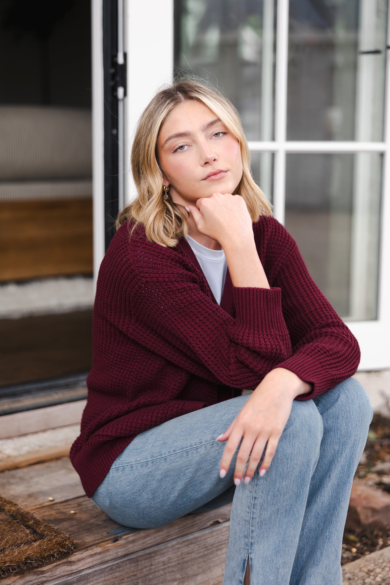 Modal sitting outdoors wearing Chunky Knit Women's Waffle Cardigan in Burgundy over white t-shirt and blue jeans