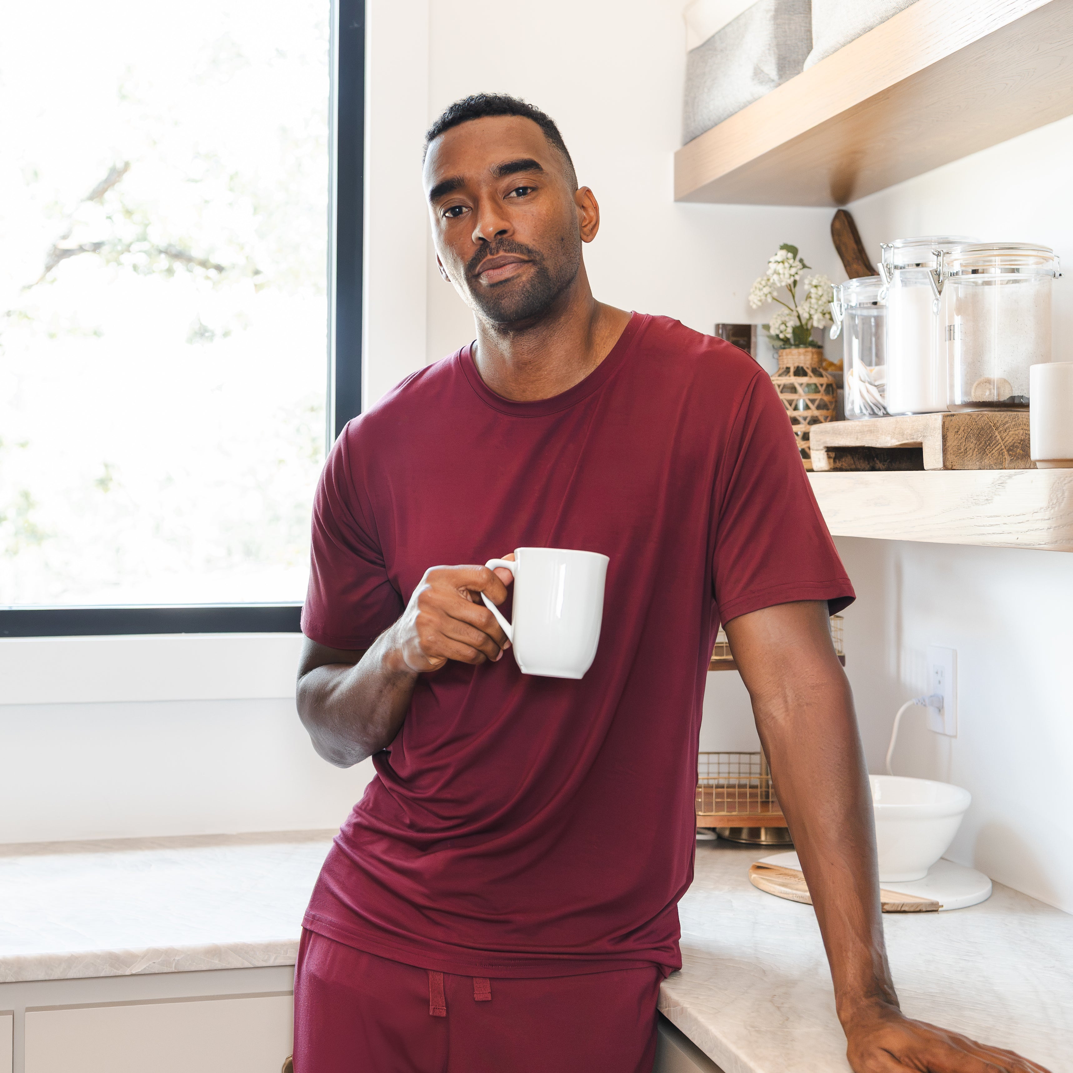 Model wearing Men's Lounge Pants in Burgundy under Men's crew neck tee in burgundy standing with a mug in a kitchen
