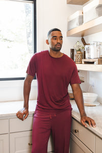 Model wearing Men's Lounge Pants in Burgundy under Men's crew neck tee in burgundy standing in a kitchen