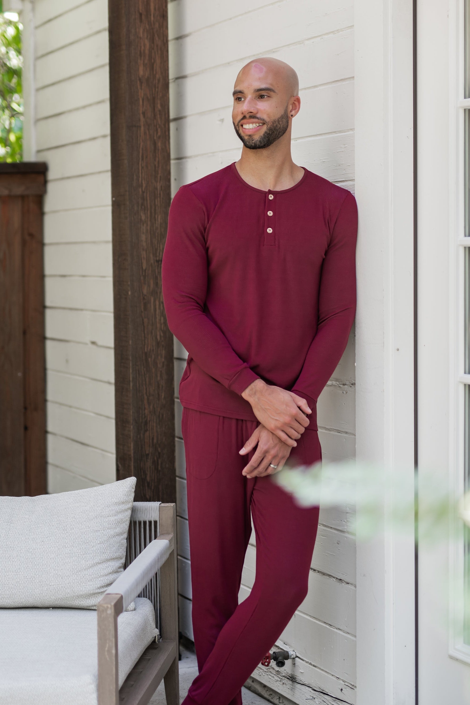 Male model leaning against a white house looking off to the side wearing the Men's Ribbed Henley Set in Burgundy