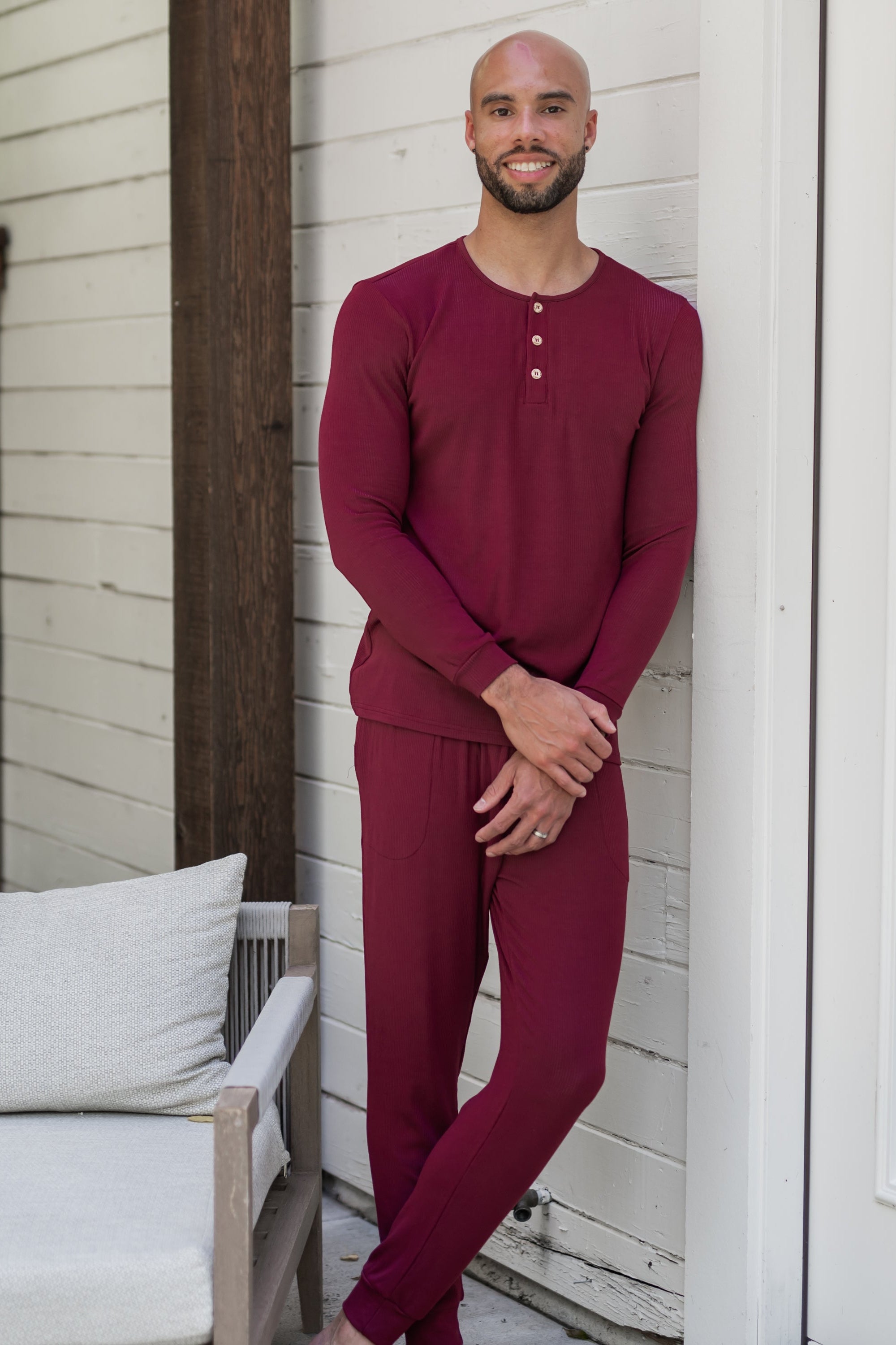 Male model leaning against a white house wearing the Men's Ribbed Henley Set in Burgundy
