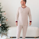 Male smiling looking off to the side standing in front of a cream ottoman and two decorated Christmas trees wearing the Men's Ribbed Henley Set in Burgundy Stripe