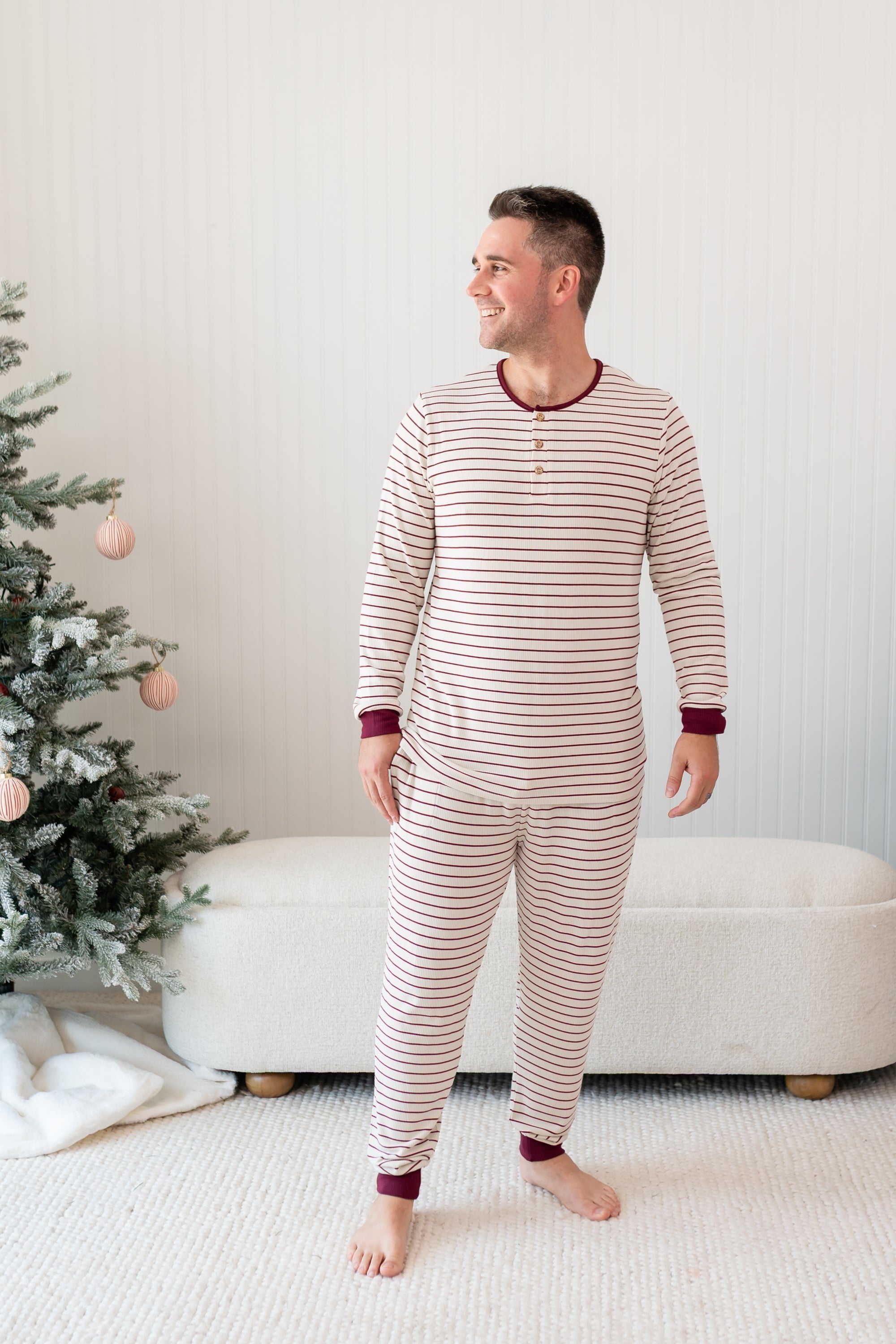 Male smiling looking off to the side standing in front of a cream ottoman and two decorated Christmas trees wearing the Men's Ribbed Henley Set in Burgundy Stripe