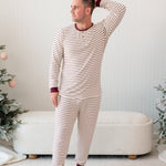Male model standing in front of a cream ottoman between two christmas trees wearing the Men's Ribbed Henley Set in Burgundy Stripe with one hand behind his head