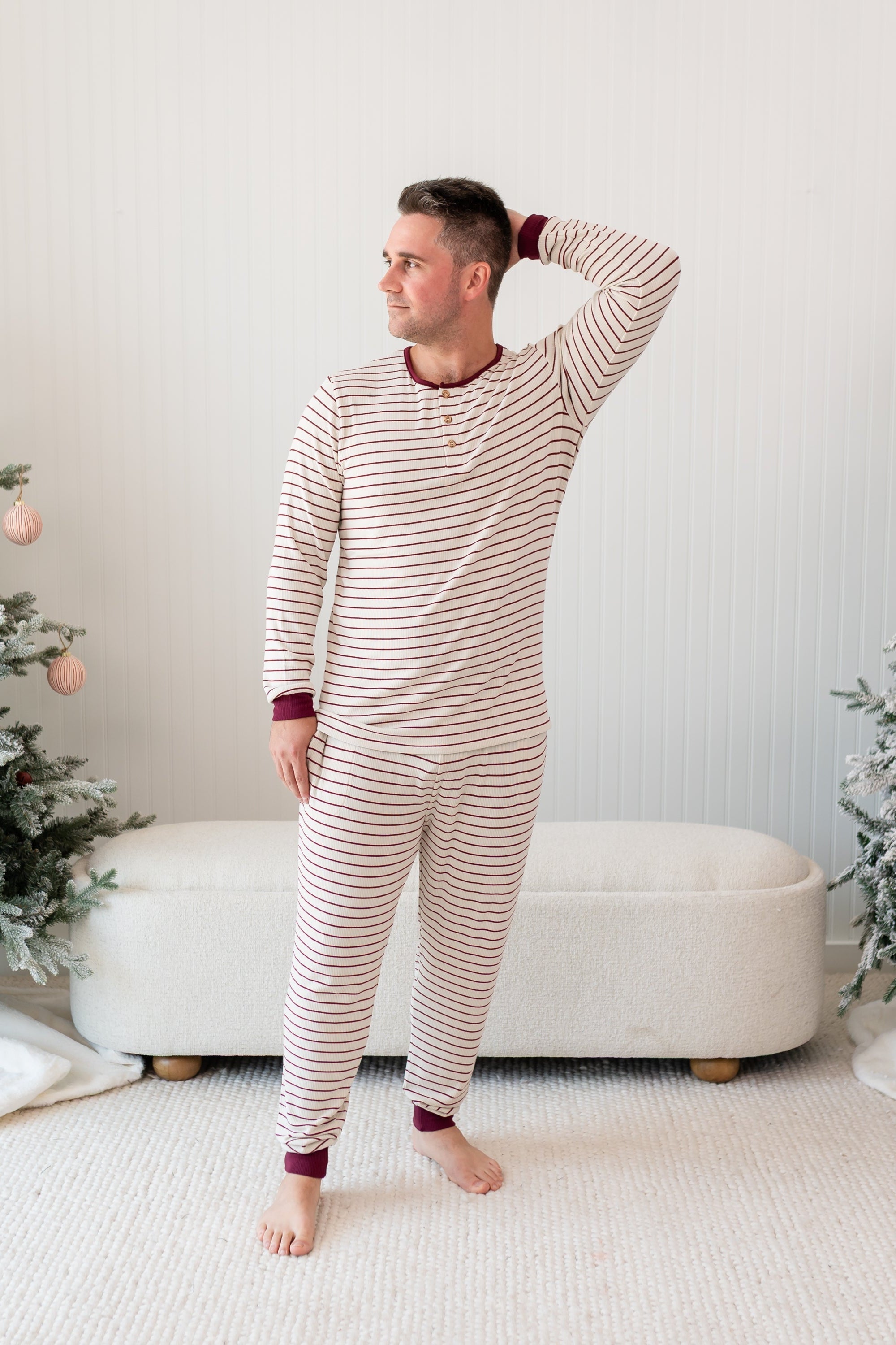 Male model standing in front of a cream ottoman between two christmas trees wearing the Men's Ribbed Henley Set in Burgundy Stripe with one hand behind his head