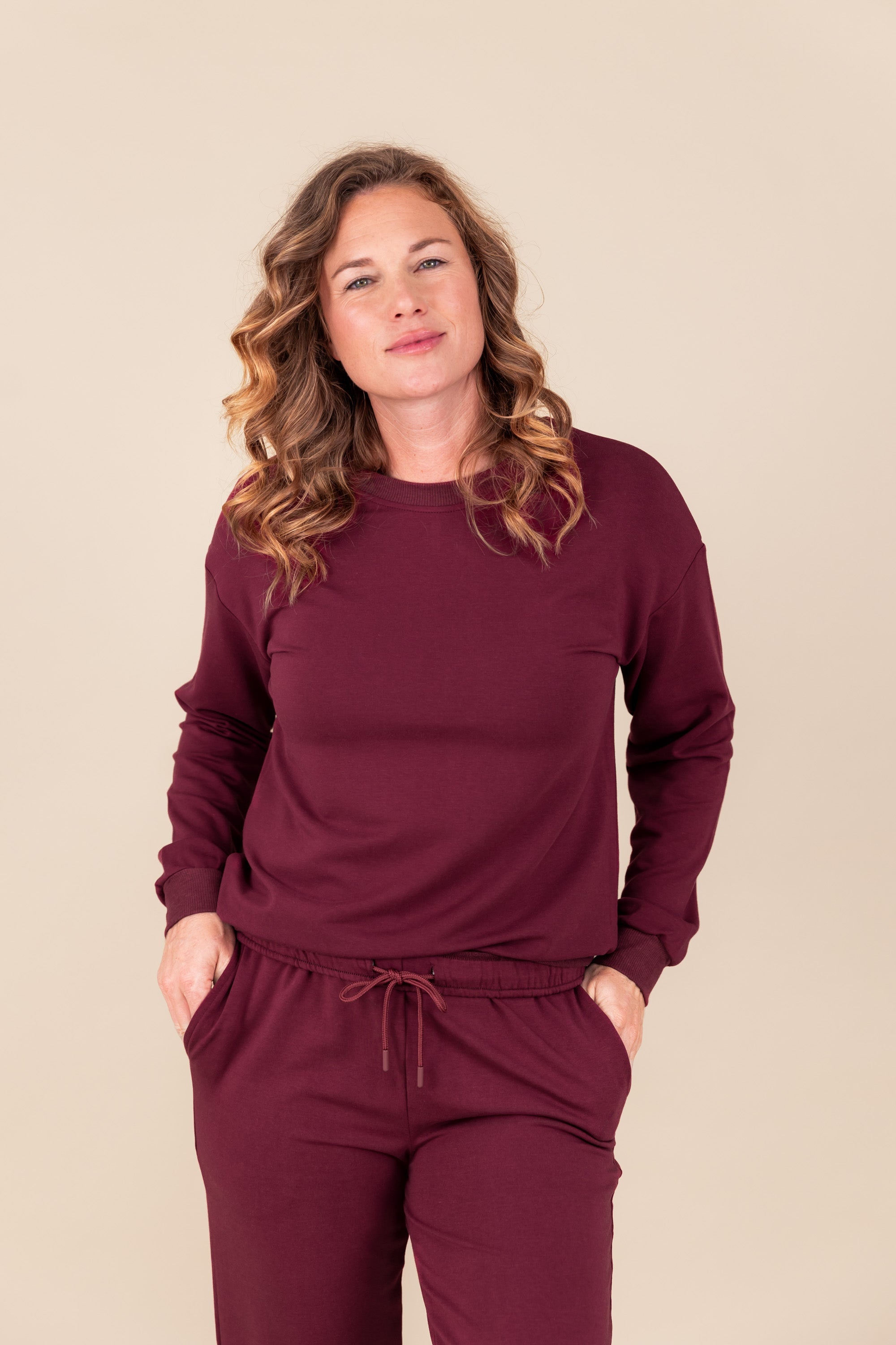 Woman smirking with both hands in her sweatpant pockets with a sweatshirt on in burgundy
