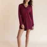 A woman modeling a long-sleeve burgundy button-up shirt dress against a solid beige studio background.