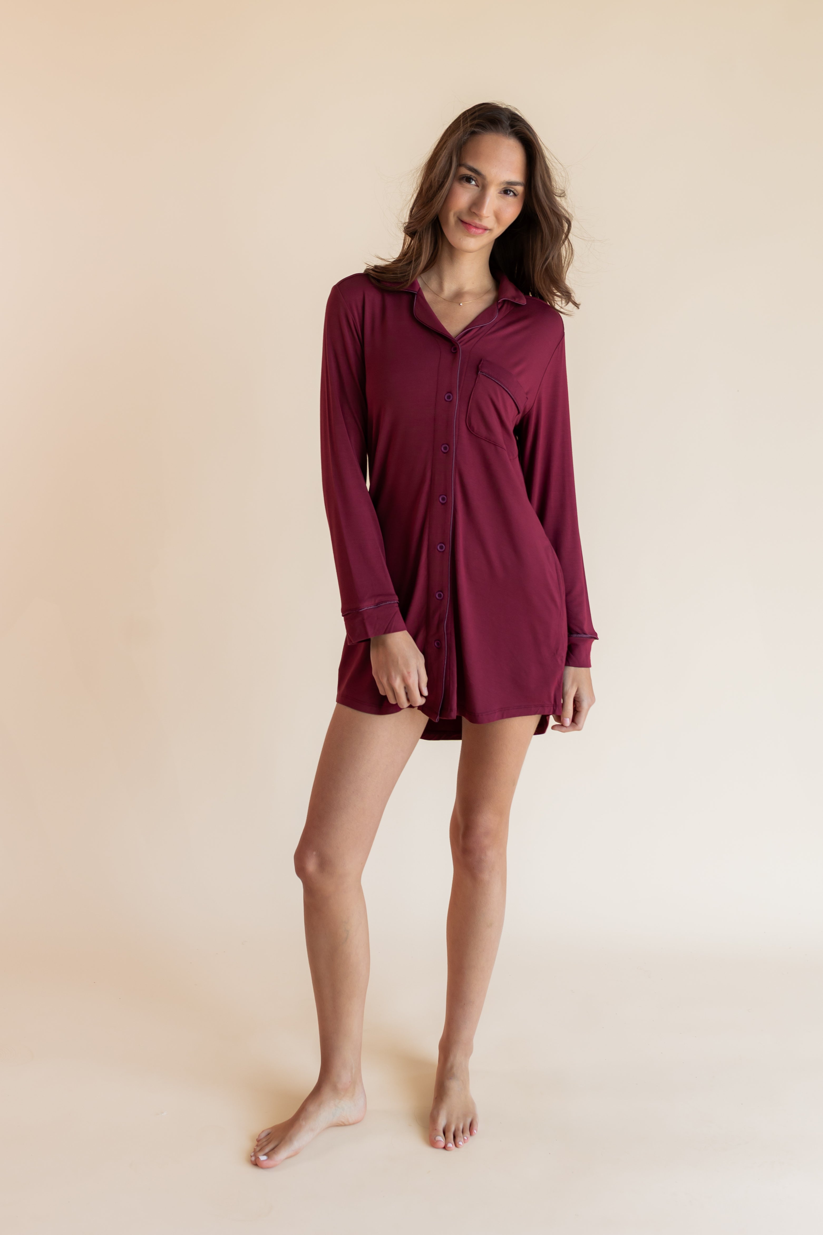 A woman modeling a long-sleeve burgundy button-up shirt dress against a solid beige studio background.