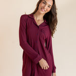 A woman modeling a long-sleeve burgundy button-up shirt dress against a solid beige studio background.