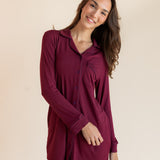 A woman modeling a long-sleeve burgundy button-up shirt dress against a solid beige studio background.