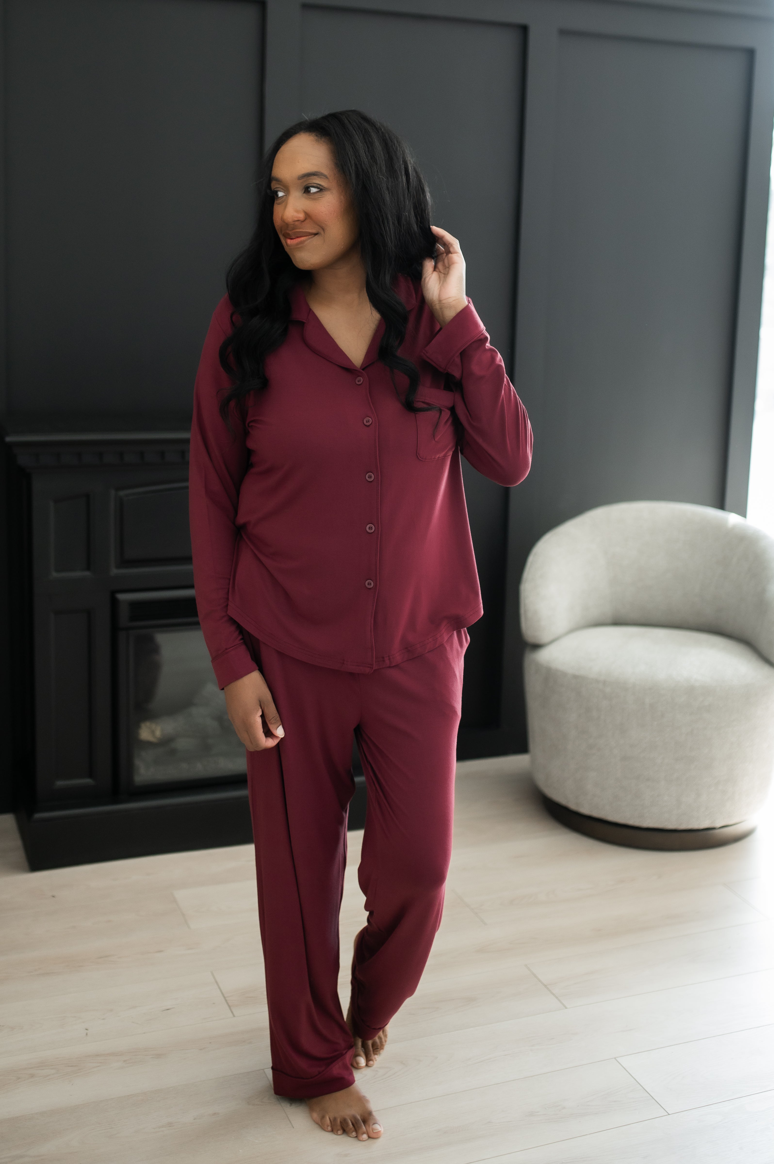 Woman standing in front of a black wall and fireplace while modeling the Bamboo Long-Sleeved Women's Pajama Set in Burgundy with Burgundy Trim