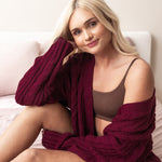 Woman wearing a burgundy cable knit bamboo cardigan and deep brown bralette sitting on a bed