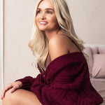 Woman wearing a burgundy knitted sweater sitting on a bed.