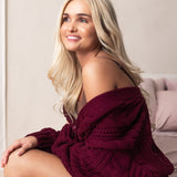 Woman wearing a burgundy knitted sweater sitting on a bed.