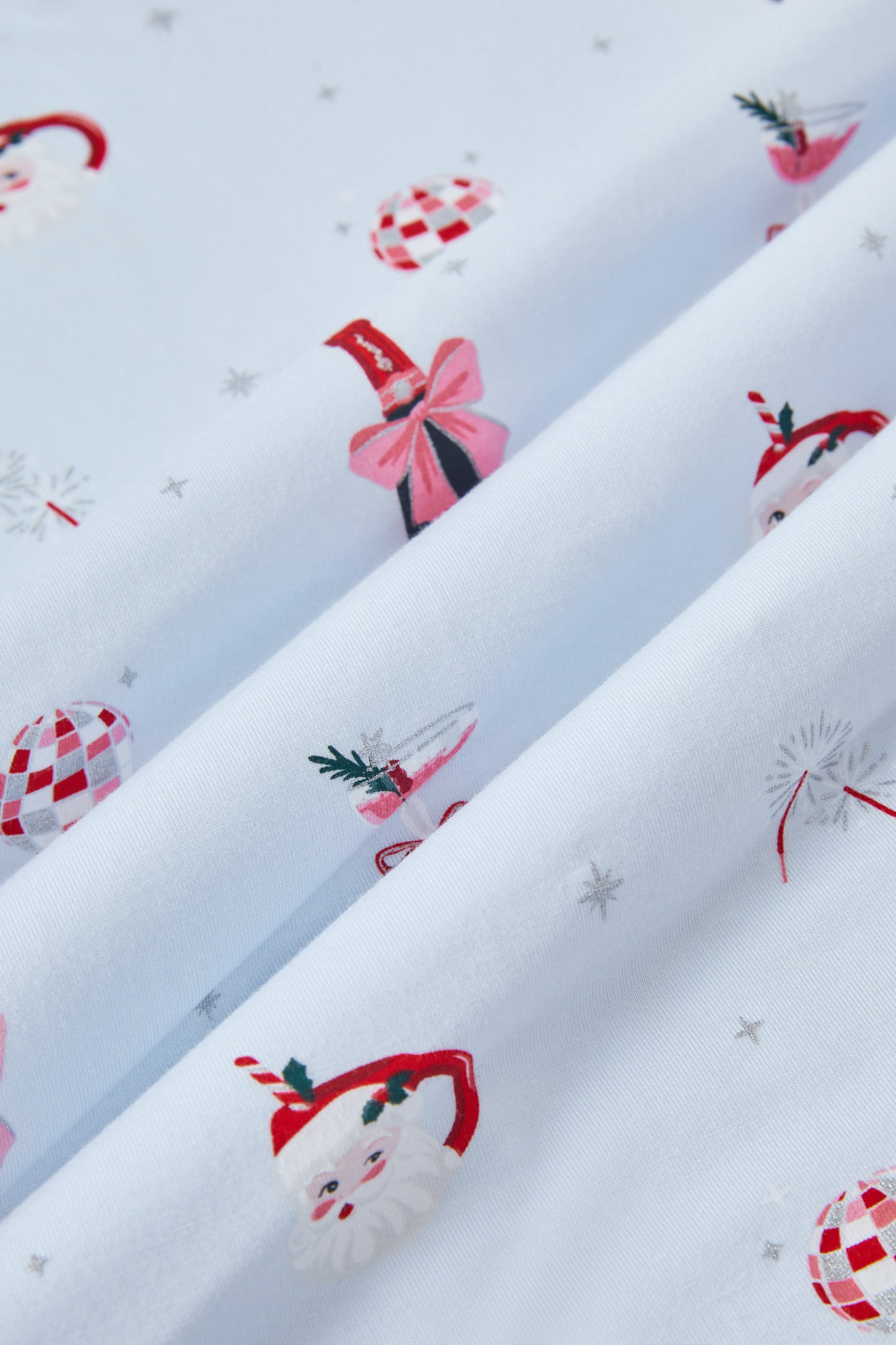 A repeating, all-over print featuring: a bottle of sparkling wine with pink packaging and a pink bow; pink bubbly in a coupe glass with a red bow tied around the stem and garnished with a sprig of rosemary and red berries; a pair of pink sparklers; a Santa Claus mug with a candy cane straw and a holly leaf; a pink-and-silver disco ball, and various metallic silver sparkles over a pale blue background color