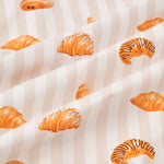 An all-over print of various types of croissants over a background of vertical stripes in white and a light beige color named Vanilla.