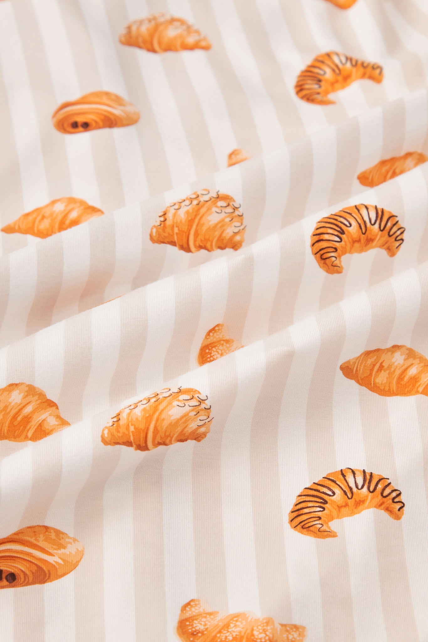 An all-over print of various types of croissants over a background of vertical stripes in white and a light beige color named Vanilla.
---1620 1
