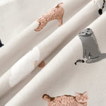 A close-up of draped fabric featuring a playful print of various cartoon-style cats in shades of gray, white, and warm tan on an off-white background.