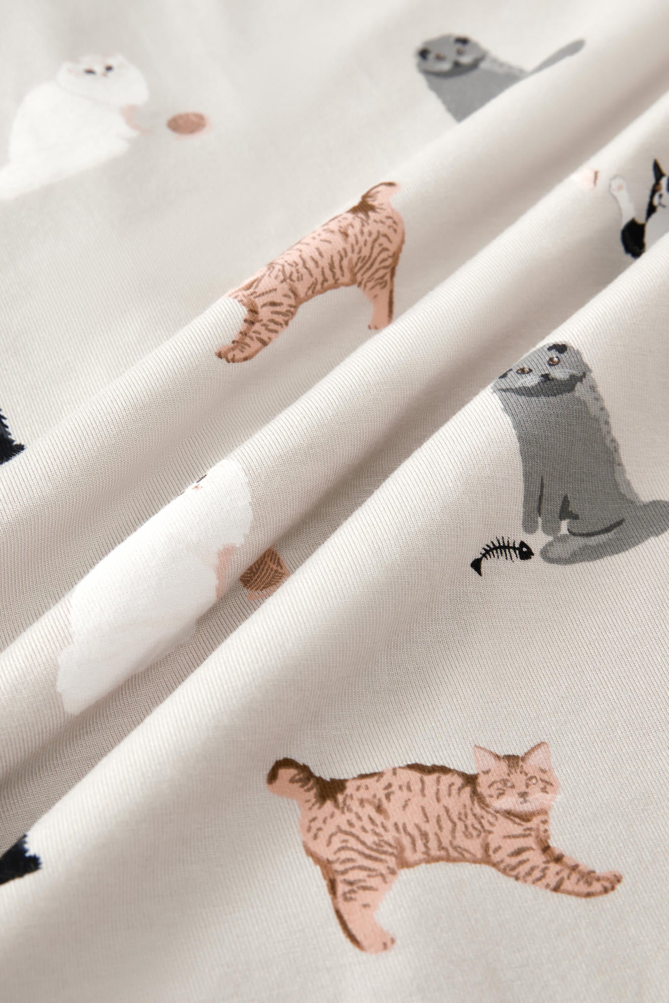 A close-up of draped fabric featuring a playful print of various cartoon-style cats in shades of gray, white, and warm tan on an off-white background.