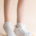 A close-up of a person's lower legs and feet, showing one ankle in a light gray ribbed ankle sock and the other in a white athletic sneaker, against a solid pale peach background.