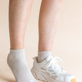 A close-up of a person's lower legs and feet, showing one ankle in a light gray ribbed ankle sock and the other in a white athletic sneaker, against a solid pale peach background.