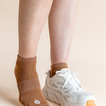 A close-up of a person's legs wearing a pair of low-cut brown knit socks and a white athletic sneaker against a solid beige background.