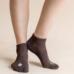 A close-up of a person's feet wearing dark brown low-cut socks with a small white geometric logo on the toes, set against a solid beige background.