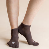 A close-up of a person's feet wearing dark brown low-cut socks with a small white geometric logo on the toes, set against a solid beige background.