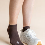 A person's legs and feet shown wearing a dark brown ankle sock on one foot and a white athletic sneaker on the other, against a plain beige studio background.