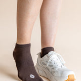 A person's legs and feet shown wearing a dark brown ankle sock on one foot and a white athletic sneaker on the other, against a plain beige studio background.