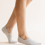 A close-up of a person's feet wearing light gray low-cut no-show socks against a plain, warm off-white background.