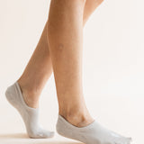 A close-up of a person's feet wearing light gray low-cut no-show socks against a plain, warm off-white background.