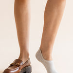 A close-up of a person's legs showing two different styles of low-cut socks: one leg wears a brown leather loafer with a chunky sole, while the other leg wears a light gray ankle sock.